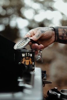 A tattooed hand is holding a can labeled with the word 'WESN', set against a blurred natural background. The scene has a rustic feel with blurred trees in the background, suggesting an outdoor setting.