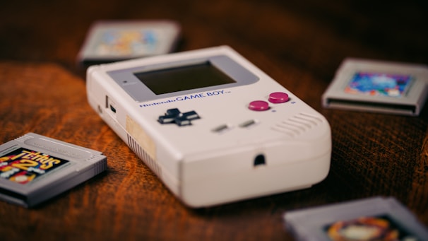 A vintage handheld gaming device is positioned prominently on a wooden surface. Surrounding the device are several gray game cartridges, each with colorful labels. The device has a monochrome display and red-colored buttons, showcasing a retro design characteristic of early portable gaming technology.