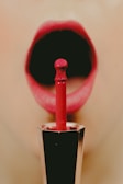 A close-up of a person's open mouth with red lipstick being applied. The focus is on the lipstick applicator and the vibrant red lips, creating an artistic and bold composition.