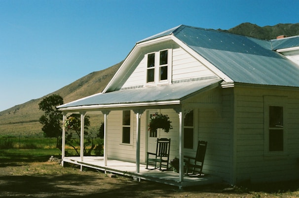 A charming white wooden house with a porch sits in a rural landscape. Two rocking chairs are placed on the porch next to a hanging flower basket. The structure features a metal roof, and the backdrop includes a grassy field and distant mountains under a clear blue sky.