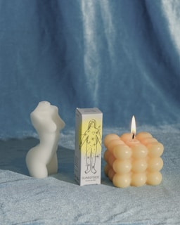 A white, sculptural candle is shaped like a torso, standing beside a box labeled 'Sunnyside' featuring an illustration of a figure with stars. Next to it, there is a lit candle with a geometric, bubble-like surface. The background is a soft, velvety blue fabric.