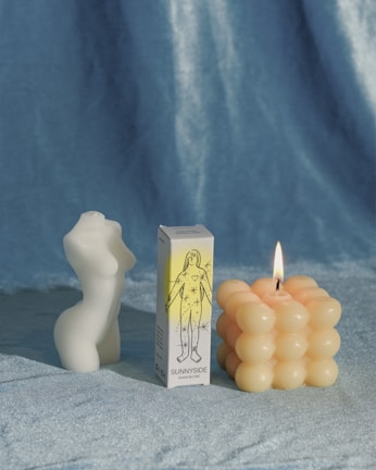 A white, sculptural candle is shaped like a torso, standing beside a box labeled 'Sunnyside' featuring an illustration of a figure with stars. Next to it, there is a lit candle with a geometric, bubble-like surface. The background is a soft, velvety blue fabric.