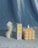 A white, sculptural candle is shaped like a torso, standing beside a box labeled 'Sunnyside' featuring an illustration of a figure with stars. Next to it, there is a lit candle with a geometric, bubble-like surface. The background is a soft, velvety blue fabric.