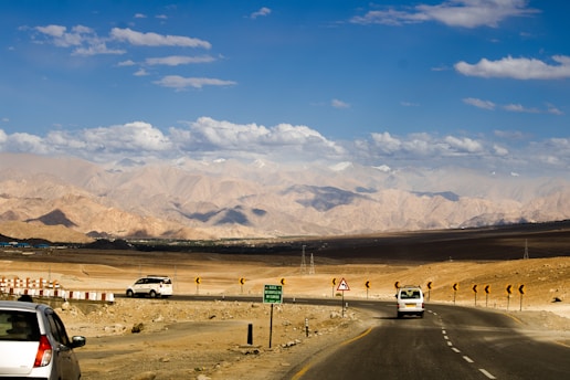 A scenic highway cuts through a barren desert landscape with rugged mountains in the distance, under a sky filled with patches of white clouds. Several vehicles travel along the road, which is lined with road signs and safety barriers.