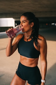 A fit woman is standing in a dimly lit space, wearing a black sports bra and shorts. She is holding a pink drink in a transparent bottle and is in the act of taking a sip. Her hair is pulled back, and she wears a watch on her left wrist.