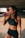 A fit woman is standing in a dimly lit space, wearing a black sports bra and shorts. She is holding a pink drink in a transparent bottle and is in the act of taking a sip. Her hair is pulled back, and she wears a watch on her left wrist.