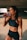 A fit woman is standing in a dimly lit space, wearing a black sports bra and shorts. She is holding a pink drink in a transparent bottle and is in the act of taking a sip. Her hair is pulled back, and she wears a watch on her left wrist.