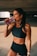 A fit woman is standing in a dimly lit space, wearing a black sports bra and shorts. She is holding a pink drink in a transparent bottle and is in the act of taking a sip. Her hair is pulled back, and she wears a watch on her left wrist.