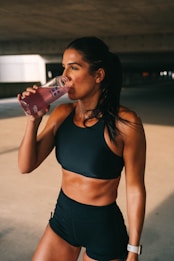 A fit woman is standing in a dimly lit space, wearing a black sports bra and shorts. She is holding a pink drink in a transparent bottle and is in the act of taking a sip. Her hair is pulled back, and she wears a watch on her left wrist.