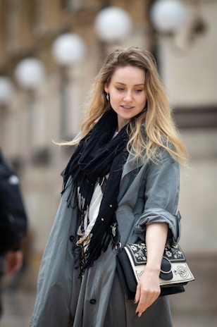 A stylish young woman wearing a beige trench coat and soft gray scarf, walking through a sunlit city street.