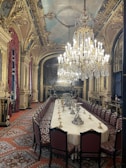 An opulent dining room featuring a long, elegantly set table surrounded by upholstered chairs. Above, large, ornate chandeliers hang, illuminating the room with a warm glow. The walls and ceiling are adorned with intricate gold detailing and classical artwork, and the carpet under the table is richly patterned.
