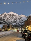 A picturesque mountain town street at dusk, flanked by snow-covered mountains in the background. String lights hang overhead, casting a warm glow on the street below. Cars are parked along the sides of the road, and a few vehicles can be seen driving down the snow-covered street. Buildings line both sides, with some people walking along the sidewalks. The scene is adorned with holiday decorations, including a lit-up Christmas tree in the distance.