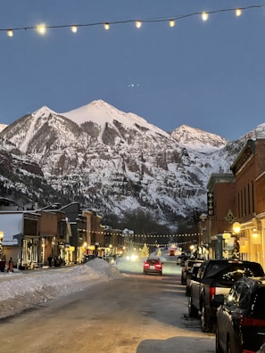 A picturesque mountain town street at dusk, flanked by snow-covered mountains in the background. String lights hang overhead, casting a warm glow on the street below. Cars are parked along the sides of the road, and a few vehicles can be seen driving down the snow-covered street. Buildings line both sides, with some people walking along the sidewalks. The scene is adorned with holiday decorations, including a lit-up Christmas tree in the distance.