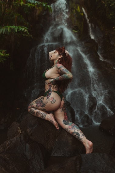 A tattooed individual poses gracefully on a rock with a backdrop of a cascading waterfall surrounded by lush greenery.