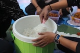 A group of people is sitting around a container of rice, with one person serving rice using a pink spatula. The container is green and placed on a surface covered with cloth. There are other food items in plastic containers nearby.