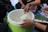 A group of people is sitting around a container of rice, with one person serving rice using a pink spatula. The container is green and placed on a surface covered with cloth. There are other food items in plastic containers nearby.