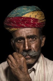 A man with a weathered face wears a vibrantly colored turban featuring blue, yellow, and red hues. He has a prominent mustache and is gazing intently at the camera. His hand, marked with a symbol, is raised thoughtfully to his chin. The background is dark, focusing attention on his facial features and attire.