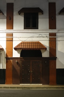 A dimly lit exterior of a building featuring a large wooden door with ornate metal handles, flanked by two lantern-style wall lights. The facade includes red brick columns and a small canopy over the door and window above. Electrical wires are visible across the front.