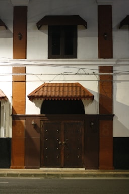 A dimly lit exterior of a building featuring a large wooden door with ornate metal handles, flanked by two lantern-style wall lights. The facade includes red brick columns and a small canopy over the door and window above. Electrical wires are visible across the front.