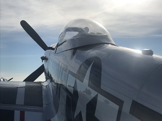 A close-up view of a vintage aircraft, focusing on the cockpit area. The sunlight reflects off the polished exterior, highlighting military insignia and details. The propeller is visible in the background against a partly cloudy sky.