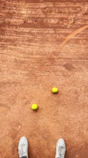 A clay tennis court with two yellow tennis balls placed near each other. A pair of white sneakers is visible at the bottom of the image, suggesting a person standing on the court.