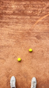 A clay tennis court with two yellow tennis balls placed near each other. A pair of white sneakers is visible at the bottom of the image, suggesting a person standing on the court.