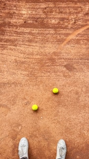 A clay tennis court with two yellow tennis balls placed near each other. A pair of white sneakers is visible at the bottom of the image, suggesting a person standing on the court.