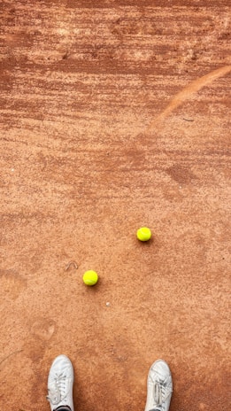 A clay tennis court with two yellow tennis balls placed near each other. A pair of white sneakers is visible at the bottom of the image, suggesting a person standing on the court.