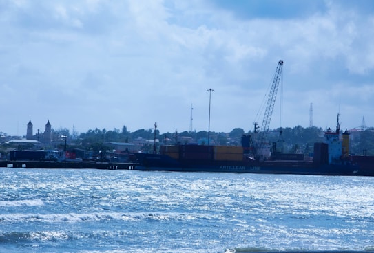 A coastal scene with a large cargo ship docked at a port. The ship has 'Antillean Line' written on its side and is equipped with cranes for loading and unloading containers. In the background, there are buildings, including one with pointed towers, suggesting a small town or cityscape. The sky is partly cloudy, and the ocean water appears choppy, reflecting the light.