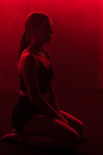 A silhouette of a person kneeling, surrounded by a deep red lighting that casts dramatic shadows. The atmosphere suggests introspection or meditation, enhanced by the stark contrast between the figure and the background.