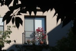 A balcony with a metal railing is adorned with potted pink-red flowers, set against a beige building wall. The window behind the balcony is covered with a light blue window shade. Dark shadows of leaves frame the outer edges of the image, suggesting a tree is nearby.