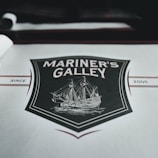 A menu cover featuring a vintage-style illustration of a sailing ship surrounded by a decorative shield. The words 'Mariner's Galley' are prominently displayed above the ship drawing. The background is predominantly gray with maroon and black accents, including the year '2005' printed on the right side.