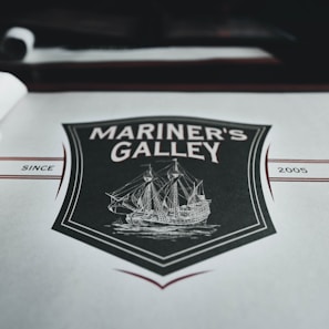 A menu cover featuring a vintage-style illustration of a sailing ship surrounded by a decorative shield. The words 'Mariner's Galley' are prominently displayed above the ship drawing. The background is predominantly gray with maroon and black accents, including the year '2005' printed on the right side.