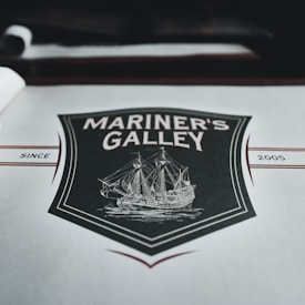 A menu cover featuring a vintage-style illustration of a sailing ship surrounded by a decorative shield. The words 'Mariner's Galley' are prominently displayed above the ship drawing. The background is predominantly gray with maroon and black accents, including the year '2005' printed on the right side.