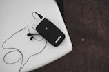 A black wireless transmitter with an attached microphone rests on a white surface. The device has 'RODELink' written on it and is connected to a cable that loops around. The background is a textured dark surface.