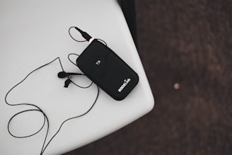 A black wireless transmitter with an attached microphone rests on a white surface. The device has 'RODELink' written on it and is connected to a cable that loops around. The background is a textured dark surface.