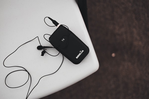 A black wireless transmitter with an attached microphone rests on a white surface. The device has 'RODELink' written on it and is connected to a cable that loops around. The background is a textured dark surface.