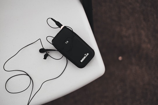 A black wireless transmitter with an attached microphone rests on a white surface. The device has 'RODELink' written on it and is connected to a cable that loops around. The background is a textured dark surface.