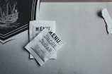 Two sugar packets labeled 'MENU' in three languages including 'SUCRE' and 'SUGAR' are placed on a table with a decorative element depicting a ship. The packaging is white with black text, and there is a tear in the paper nearby.