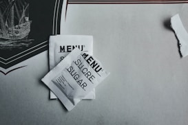Two sugar packets labeled 'MENU' in three languages including 'SUCRE' and 'SUGAR' are placed on a table with a decorative element depicting a ship. The packaging is white with black text, and there is a tear in the paper nearby.