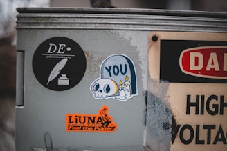 A metal surface is covered with several stickers. One black circular sticker features a feather and ink logo with the text '@dylanedwardspoetry'. Another sticker depicts a cartoon skull with candles and the word 'YOU'. An orange sticker reads 'LiUNA Local 1208 Feel the Power' with a graphic design. Part of a high voltage warning sign is also visible.