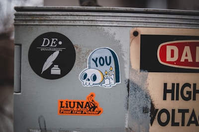 A metal surface is covered with several stickers. One black circular sticker features a feather and ink logo with the text '@dylanedwardspoetry'. Another sticker depicts a cartoon skull with candles and the word 'YOU'. An orange sticker reads 'LiUNA Local 1208 Feel the Power' with a graphic design. Part of a high voltage warning sign is also visible.