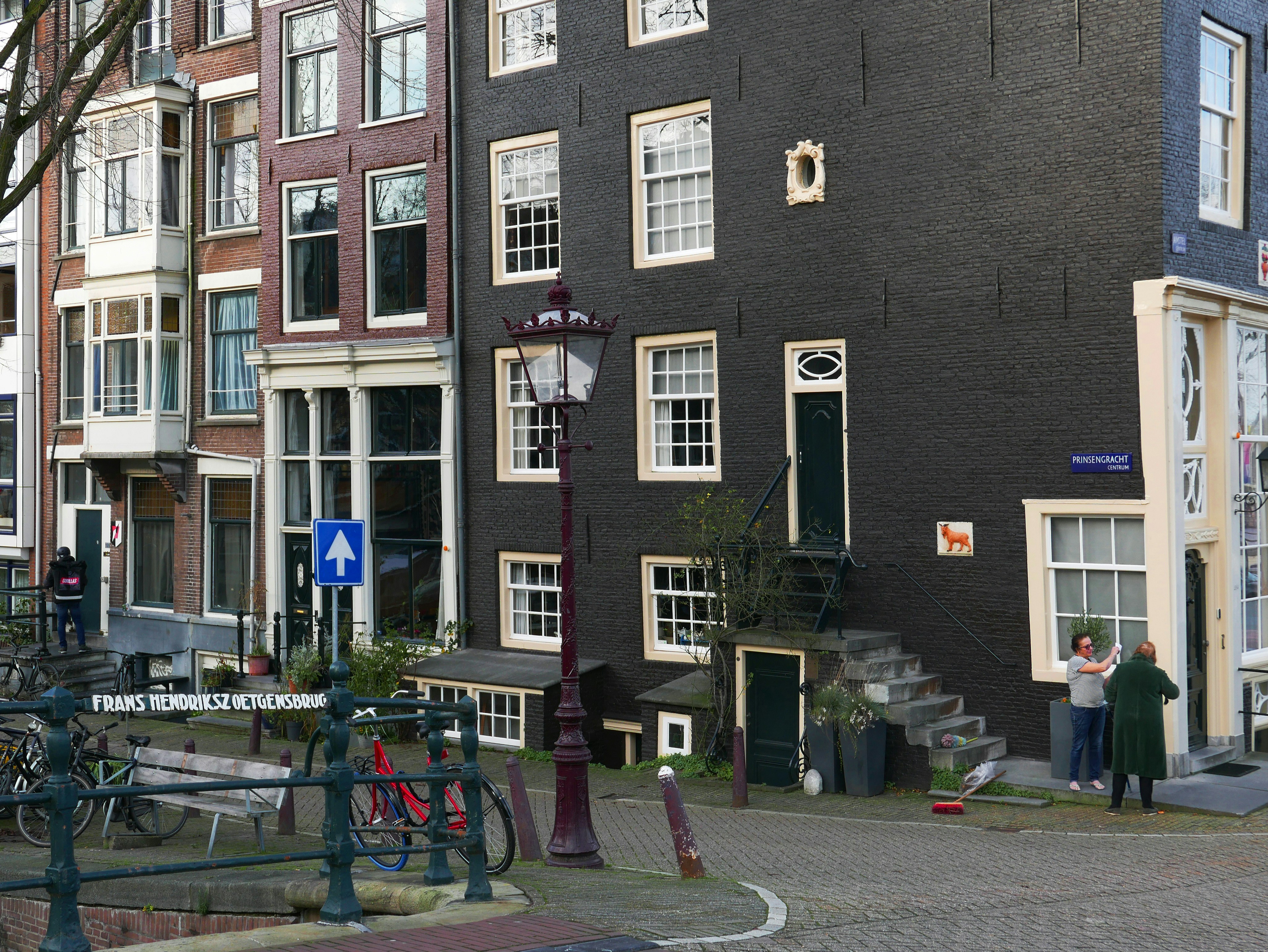 a group of people standing in front of a building, Street corner in Amsterdam old city, of the Prinsengracht street and along Amstel river. This is old architecture of mansion houses of the 17th century. The facades are brick and later painted in black at the corner. The original old canal houses had windows with rather small glasses in it, like you see here. Urban street view photography - Free photo of The Netherlands by Fons Heijnsbroek.