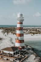 A red and white striped lighthouse stands prominently by the coastline, surrounded by a sandy beach and a modern building with outdoor seating. In the background, the sea stretches out to the horizon, bordered by palm trees. The sky is lightly clouded, adding to the coastal atmosphere.