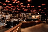 A spacious and modern coffee shop interior with an industrial-chic design. Warm lighting emanates from the grid-like ceiling, casting a cozy glow over the wooden counters and metallic coffee machines. People, wearing face masks, are seen working and enjoying their drinks in the background, adding a lively yet relaxed atmosphere.
