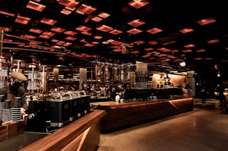 A spacious and modern coffee shop interior with an industrial-chic design. Warm lighting emanates from the grid-like ceiling, casting a cozy glow over the wooden counters and metallic coffee machines. People, wearing face masks, are seen working and enjoying their drinks in the background, adding a lively yet relaxed atmosphere.