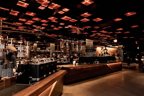 A spacious and modern coffee shop interior with an industrial-chic design. Warm lighting emanates from the grid-like ceiling, casting a cozy glow over the wooden counters and metallic coffee machines. People, wearing face masks, are seen working and enjoying their drinks in the background, adding a lively yet relaxed atmosphere.
