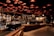 A spacious and modern coffee shop interior with an industrial-chic design. Warm lighting emanates from the grid-like ceiling, casting a cozy glow over the wooden counters and metallic coffee machines. People, wearing face masks, are seen working and enjoying their drinks in the background, adding a lively yet relaxed atmosphere.