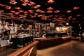 A spacious and modern coffee shop interior with an industrial-chic design. Warm lighting emanates from the grid-like ceiling, casting a cozy glow over the wooden counters and metallic coffee machines. People, wearing face masks, are seen working and enjoying their drinks in the background, adding a lively yet relaxed atmosphere.