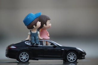 Two small figurines, a boy wearing a blue cap and overalls and a girl in a pink dress, are sitting on top of a miniature black car. The setting is a blurred, neutral background, emphasizing the playful and imaginative scene.